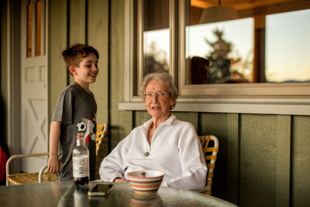 A child standing beside a smiling older woman at a porch table, family moments dementia care in Wellington protects