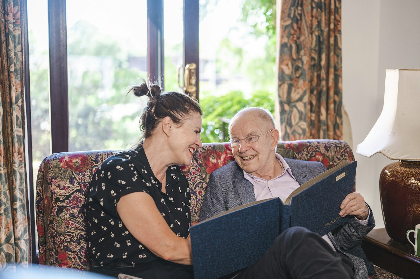 A companion and an older man laughing together over a photo album at home